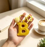 Close-up of the letter pom pom keychain showing stitched yellow PU leather letter and faux fur pom pom.