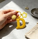 Close-up of the Letter D keychain showing padded sunny yellow PU leather and soft faux fur pom pom.