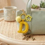 Close-up of the Letter D keychain showing padded sunny yellow PU leather and soft faux fur pom pom.